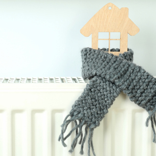 Een hout huisje met een sjaal om staat op de verwarming. De foto wordt gebruikt voor het artikel '6 tips Om Energie Te Besparen' 