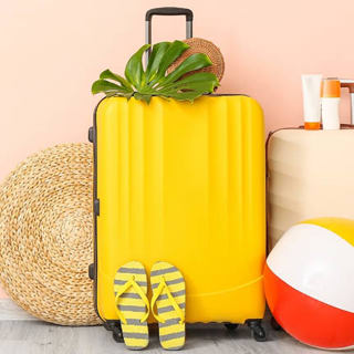 Twee vrolijke gele vakantiekoffers staan naast elkaar met links een rieten mand/tas en ervoor een strandbal en slippers. De achtergrond van de foto is perzikroze.
